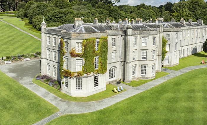 A large historic mansion with ivy-covered stone walls, surrounded by neatly manicured lawns and gardens under a bright, sunny sky